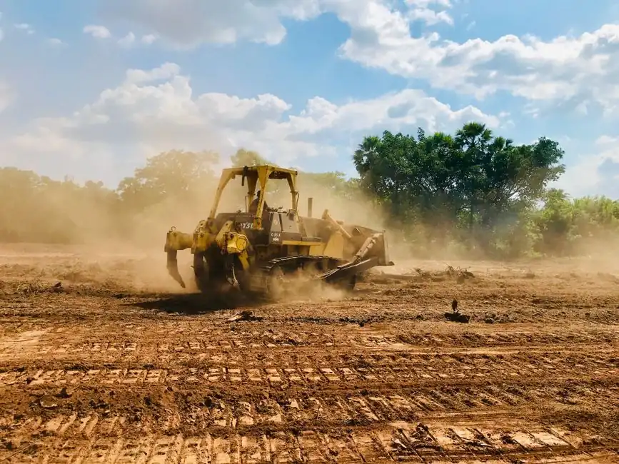 A powerful bulldozer works on a dusty, dirt field under a bright blue sky with cloud cover and trees in the background.