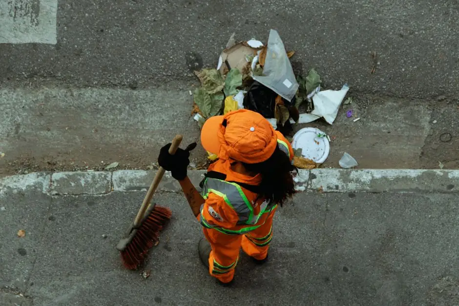 Top view of a street cleaner wearing orange uniform sweeping debris on a city sidewalk.